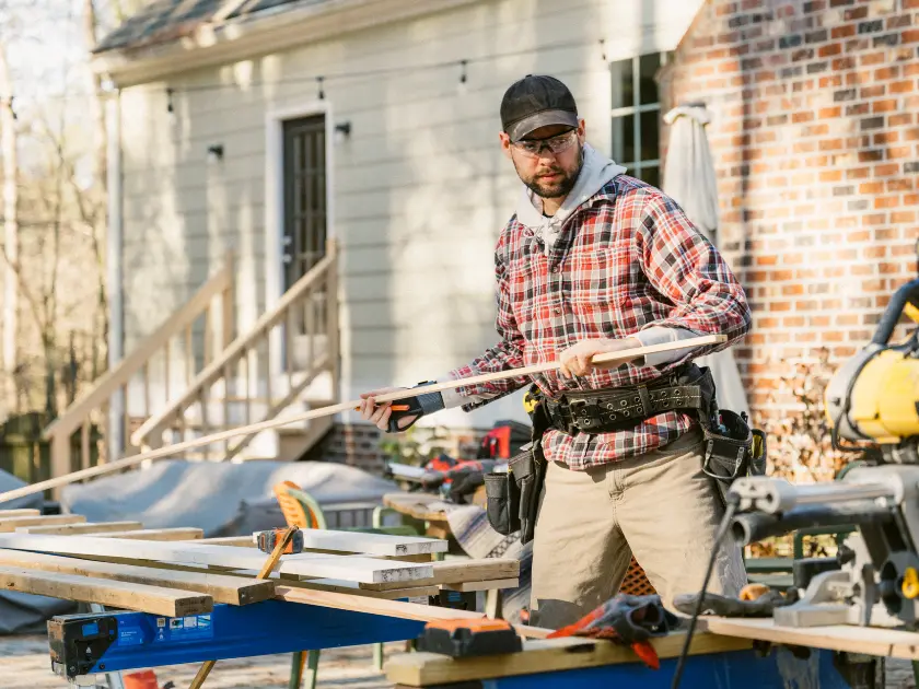 Contractor in a tool belt handling lumber at an outdoor saw station beside a house