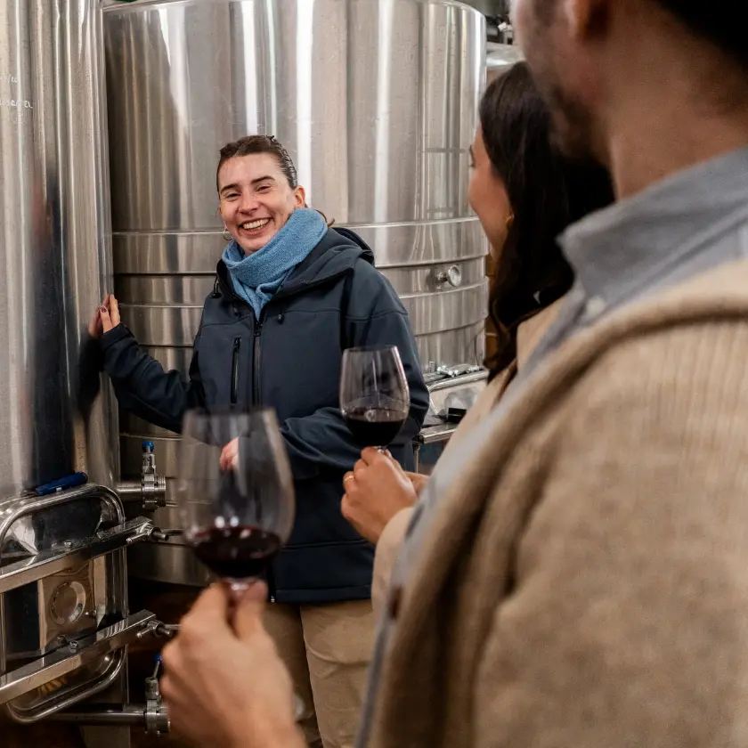 Winery host talking with visitors holding glasses of wine beside steel fermentation tanks