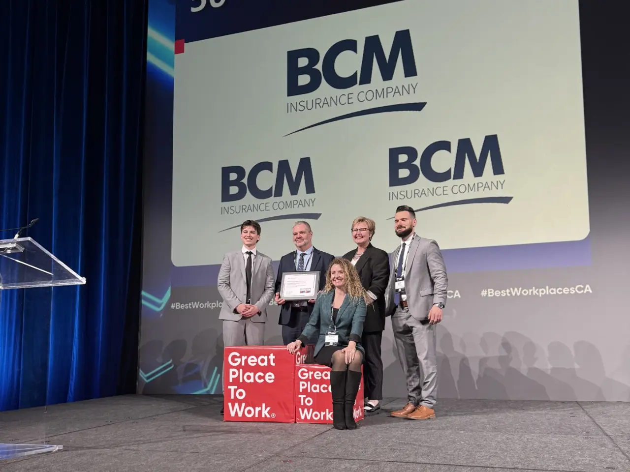 Five BCM Insurance staff on stage holding a framed certificate at a Great Place to Work event